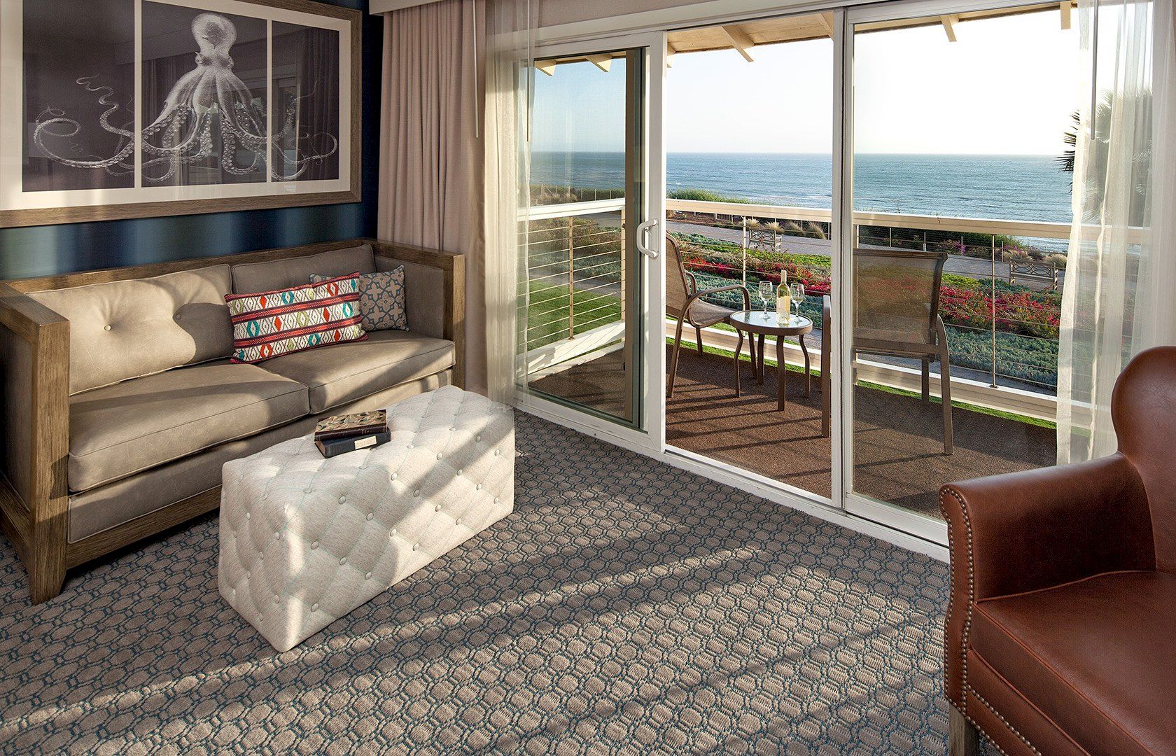 A cozy living room with a view: beige sofa, matching ottoman, artwork, and a sliding glass door opening to a balcony with ocean scenery, sunny and inviting.