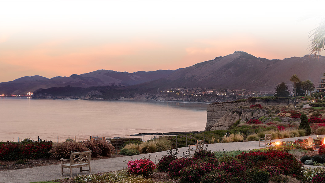 A seaside resort at sunset with calm water, mountain hills, palm trees, and landscaped gardens lit by evening lights.