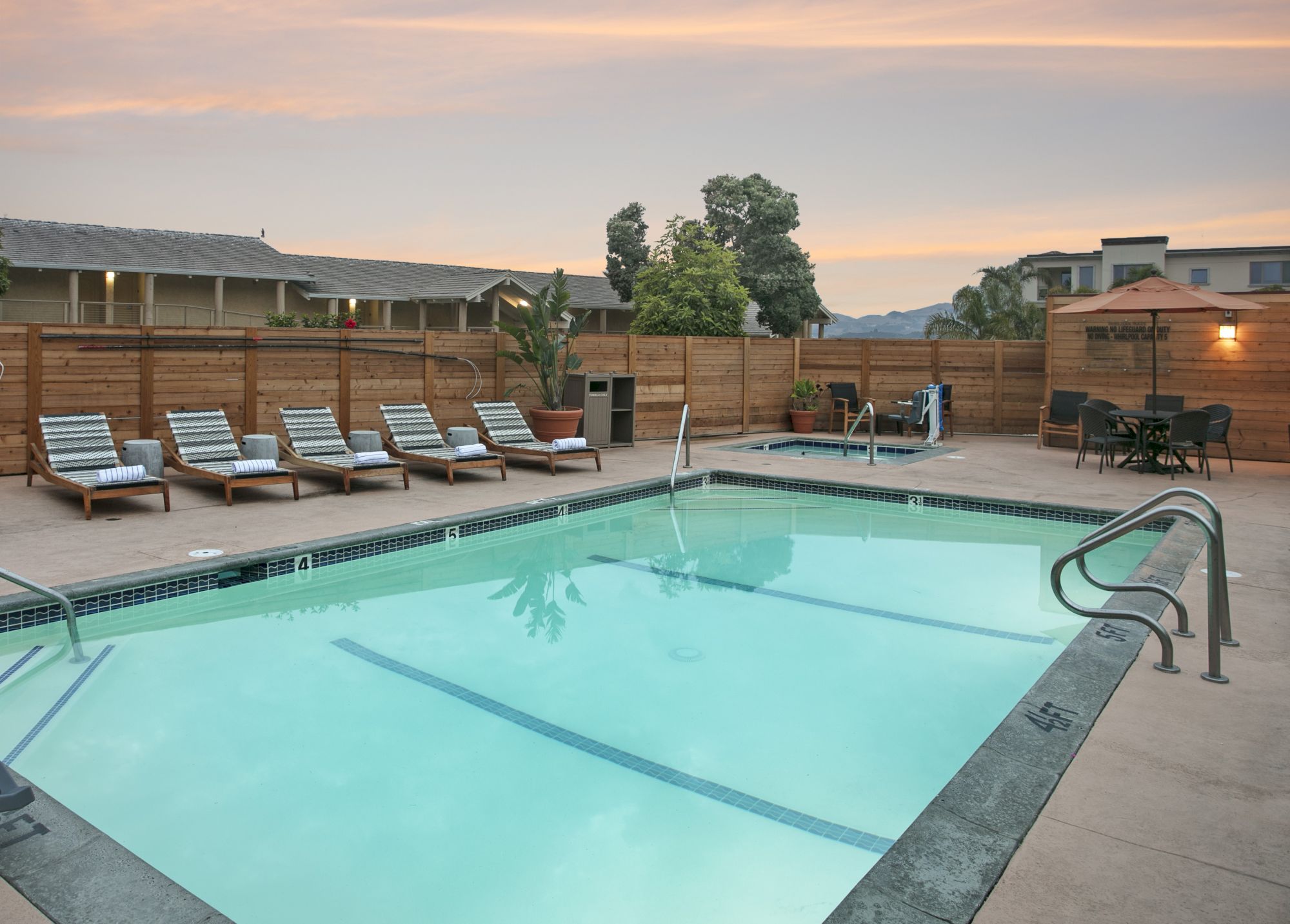 Outdoor resort pool at sunset with loungers, potted plants, and a wooden fence around the patio.
