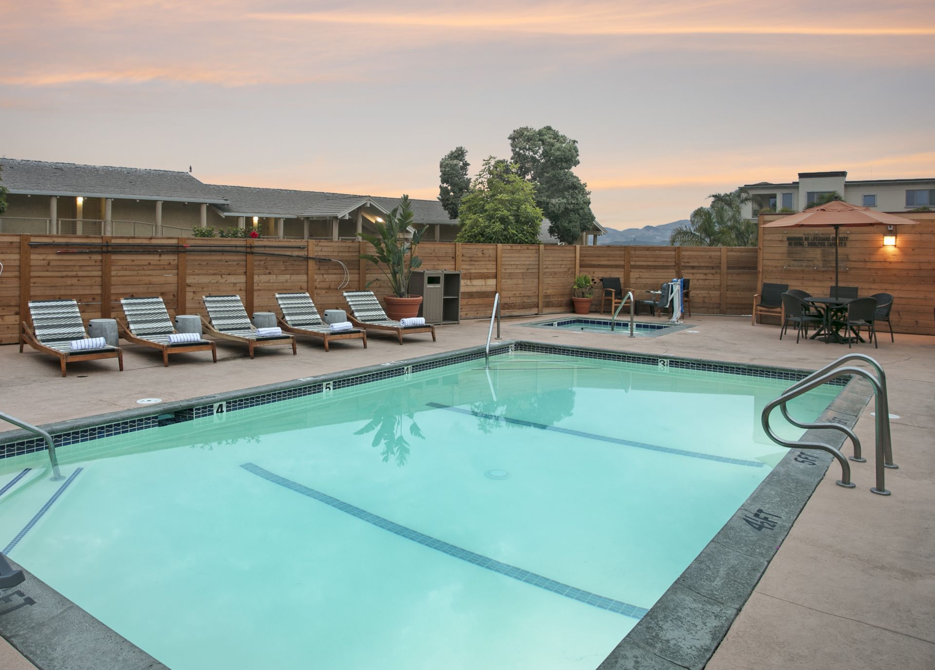 A sunny outdoor pool with lounge chairs, wooden fence, plants, and a patio at dusk&mdash;relax and swim.