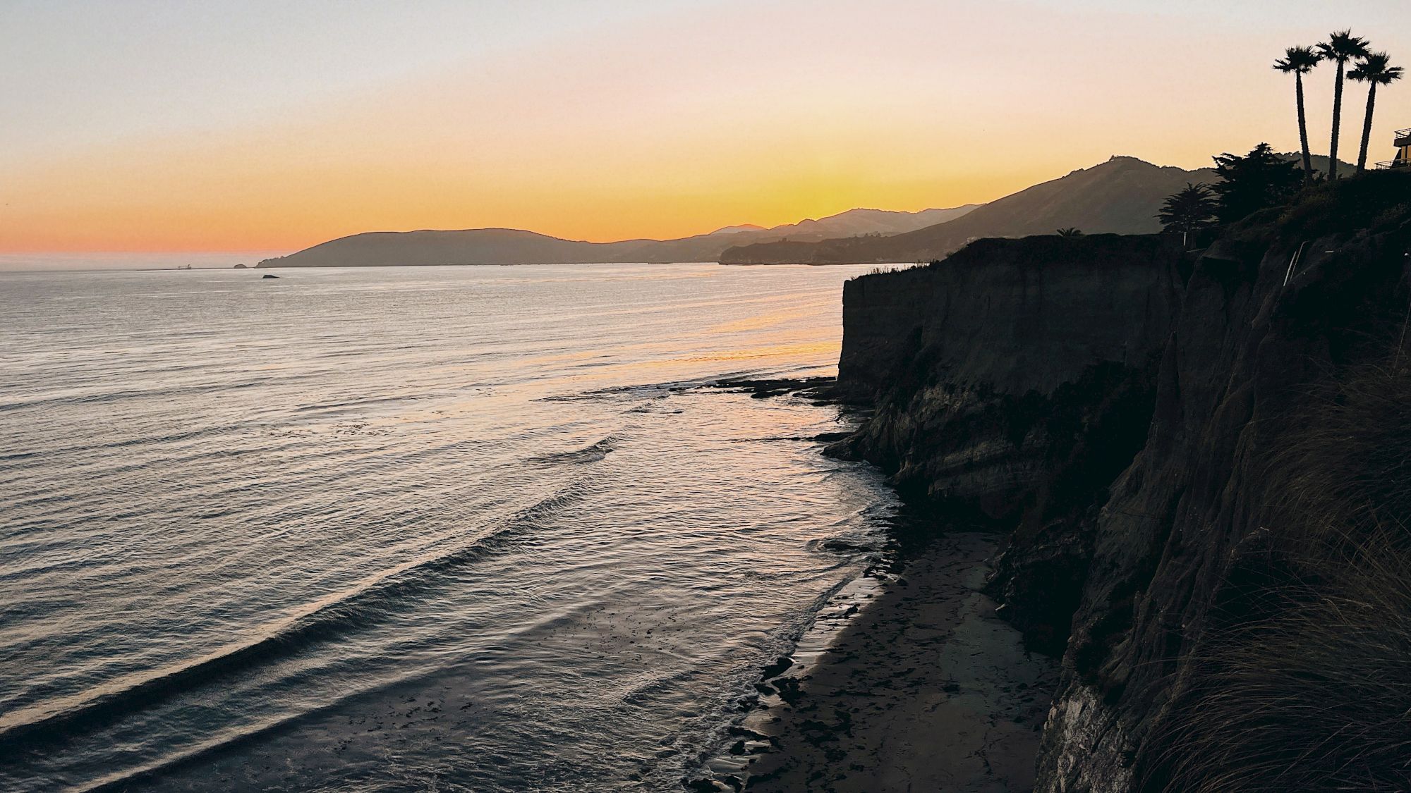 Golden sunset over a calm ocean, sandy beach, dark cliffs, and tall palm trees along the shoreline.