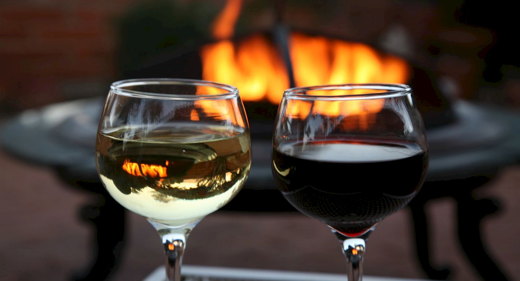 Two wine glasses, white and red, in front of a blazing fire, sharing a cozy outdoor moment.