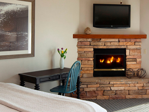 A cozy hotel-like room with a stone fireplace, mantel, and a flat-screen TV above; a desk with a vase of flowers and a blue chair, plus a bed in the foreground.