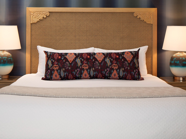 A neatly made bed with a woven headboard, two matching lamps, and a decorative dark patterned bolster pillow centered on crisp white bedding.
