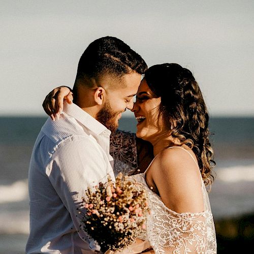 A bride and groom kiss near the ocean, holding a bouquet, smiling in wedding attire.
