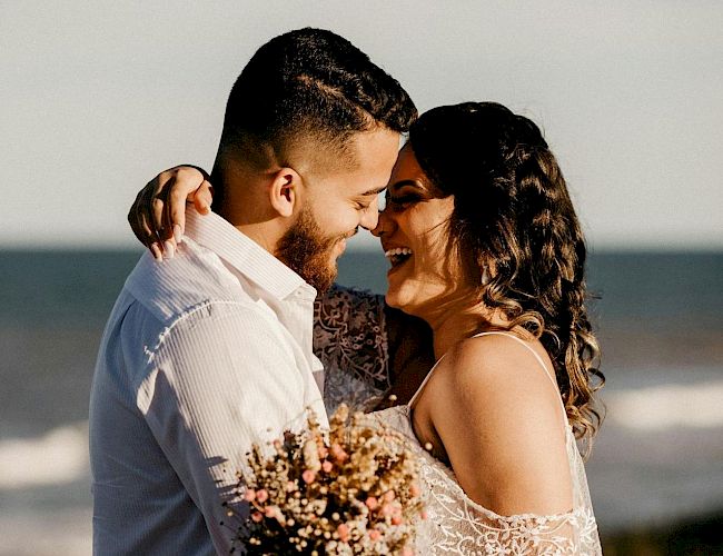 A bride and groom kiss near the ocean, holding a bouquet, smiling in wedding attire.