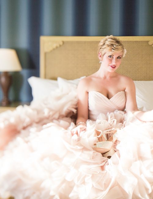 A blonde bride in a white dress lounges on a bed, holding a champagne flute in a hotel suite.