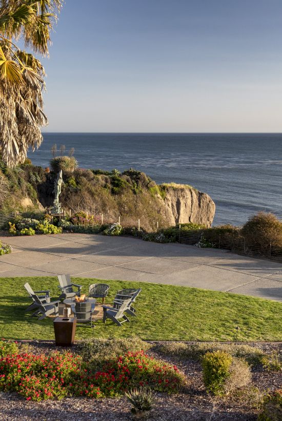 A seaside garden patio with palm trees, flowers, and a view of the ocean and rocky coastline at sunset.