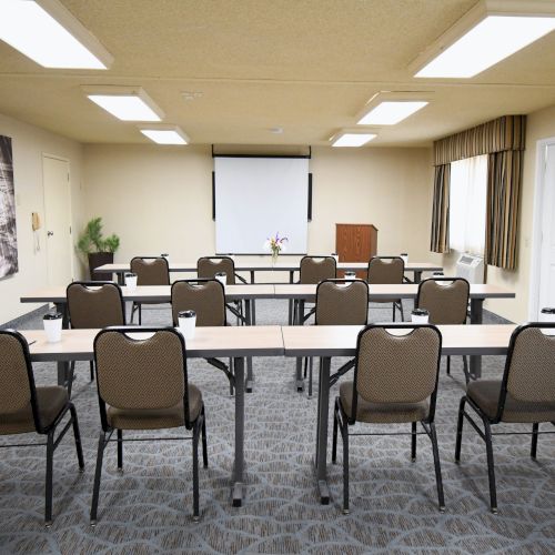 A bright meeting room with rows of chairs, tables, curtains, and a projector screen for presentations.