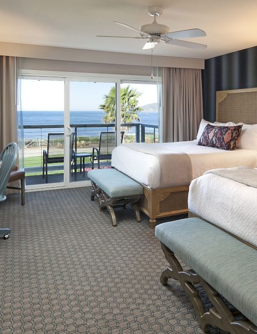 A bright hotel room with two beds, lamps, a dresser, and a window showing the ocean view outside.