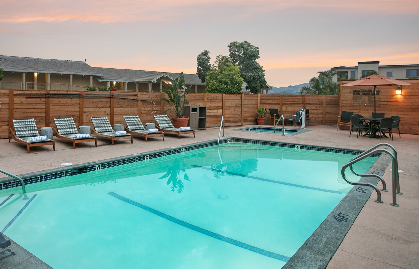 A clean outdoor pool area with turquoise water, sun loungers along the deck, a wooden fence, and a small dining setup in the corner at sunset.