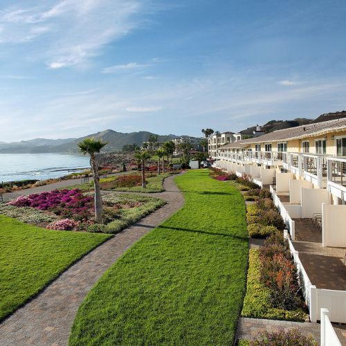 A seaside hotel corridor overlooks a manicured garden path, bright flowers, and the ocean under a blue sky.