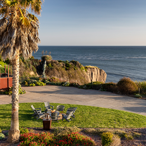 A coastal resort garden with palm trees, a stone path and patio table, overlooking cliffs and the blue ocean at sunset.