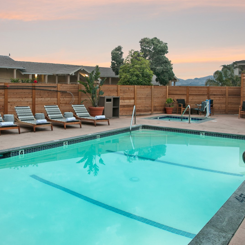 A backyard courtyard pool with sun loungers, potted plants, and a wooden fence at sunset.