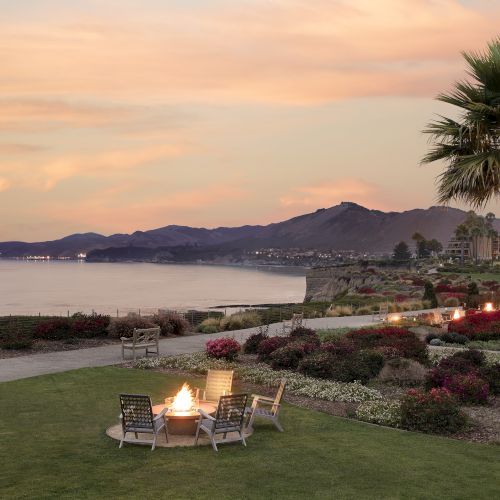 A palm-lined seaside garden patio at sunset, with chairs by a firepit, calm bay waters, and distant mountains.