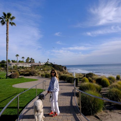 A smiling woman walks her white dog along a seaside path with palm trees, while the ocean and cloudy sky stretch behind them.