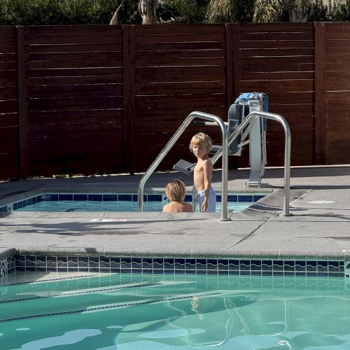 A little boy stands at a pool ladder while another child swims nearby, in an outdoor backyard with palm trees behind.