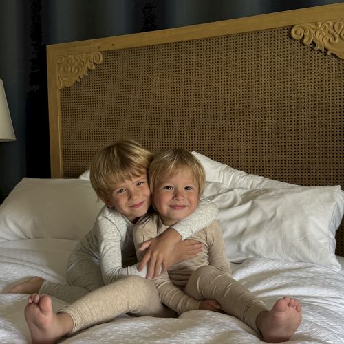 Two blond boys cuddle on a white bed, smiling in front of a decorative wooden headboard.