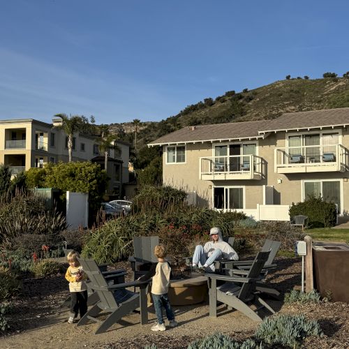 A sunny backyard patio with three people relaxing in lounge chairs, landscaped plants and flowers, and hillside homes under a blue sky.