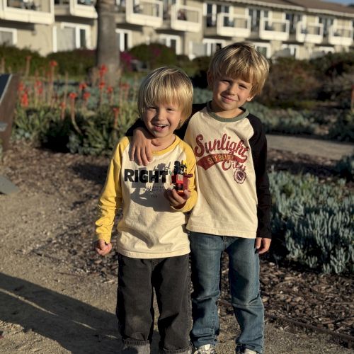 Two little boys smile in a garden courtyard, holding a colorful toy brick as warm sunlight shines on them.
