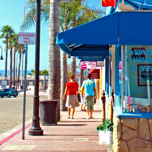 A sunny sidewalk by palm trees, two people walking past a colorful shop sign advertising an award-winning, &ldquo;open&rdquo; hot dog stand.