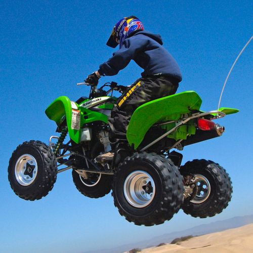 A rider in a helmet launches a green Kawasaki quad ATV off a dune against a clear blue sky.