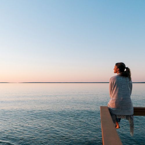 A woman stands by the ocean at sunset, looking out over calm water from a wooden deck.