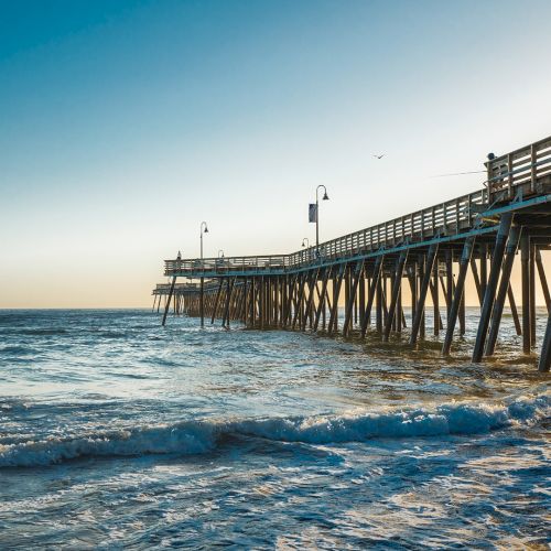 A long wooden pier on stilts stretches over the ocean as waves roll in at sunset.