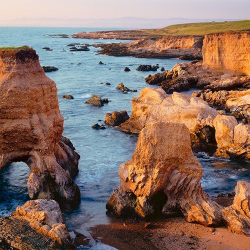 Sunlit ocean waves crash against jagged orange cliffs and boulders along a rugged coastline at golden hour.