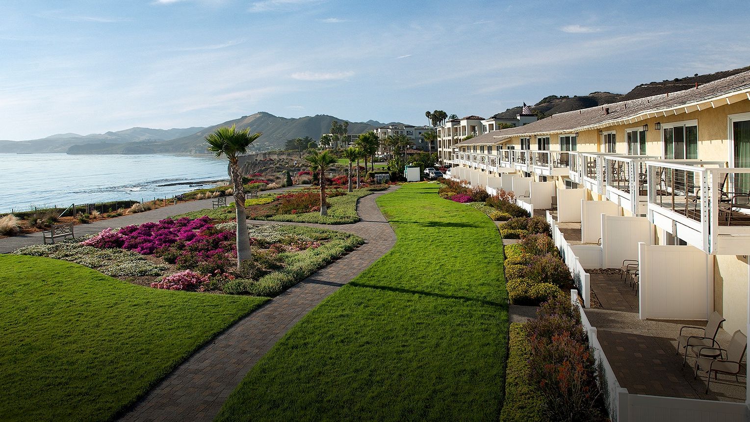 An oceanfront resort with manicured green lawns, colorful flower beds, and a row of rooms along the seaside, under a blue sky.