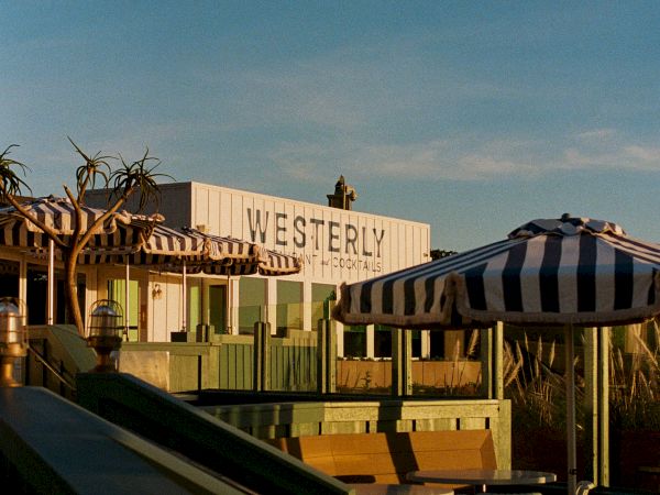 A sunny motel terrace with the &ldquo;WESTERLY&rdquo; sign, striped awnings, outdoor seating, and a clear blue sky above.