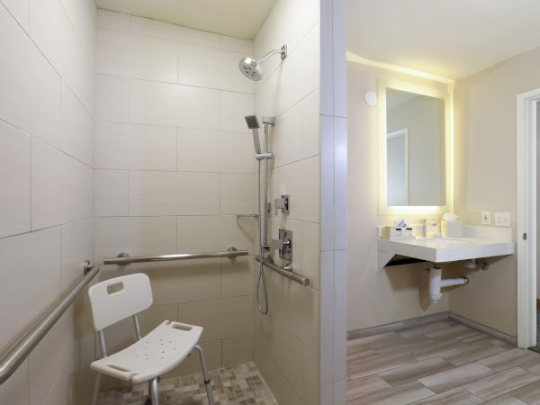 A bright accessible bathroom showing a walk-in shower with handheld showerhead, grab bars, shower chair, and a lighted vanity sink.
