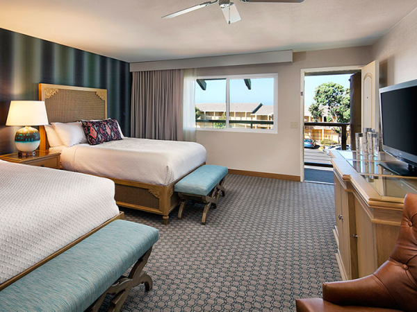 A bright hotel room with two beds, a TV stand, large window, and outdoor balcony view.