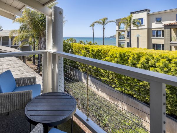 A seaside resort balcony with blue cushions, palms, lush hedges, and a view of the ocean beyond white buildings.