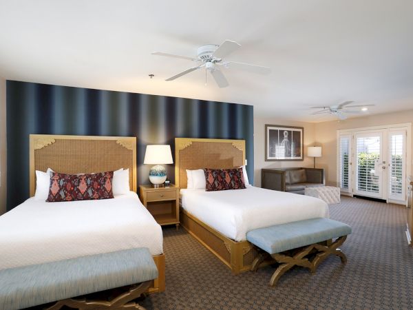A bright hotel room with two beds, bedside lamps, wall art, and a ceiling fan above.
