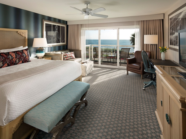 A luxury hotel room with a king bed, ocean-view balcony doors, armchairs, desk TV, and a ceiling fan overhead.