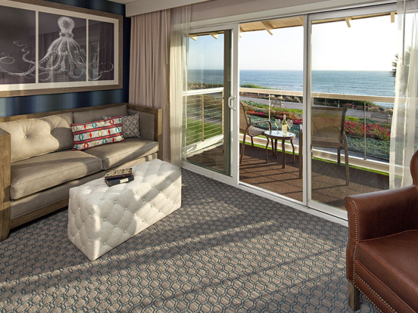 A cozy hotel room with a sofa and ottoman, large sliding doors, and a balcony view of the ocean and flowers.