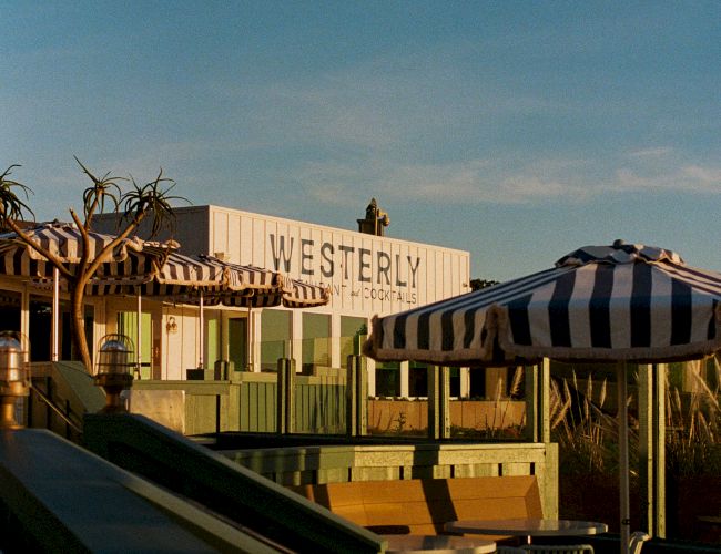 A sunny patio view of the Westerly Inn sign, cabanas and striped umbrellas against a cloudy blue sky.