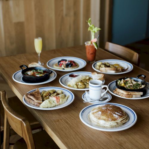 A brunch spread on a wooden table with omelets, bread, potato sides, pancakes, coffee, and a mimosa-like drink.