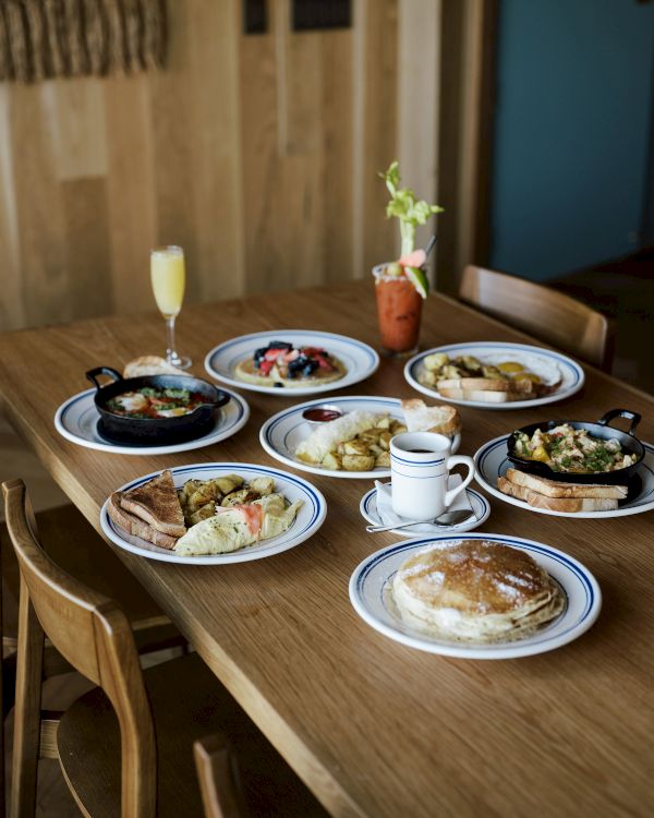 A brunch spread on a wooden table with omelets, bread, potato sides, pancakes, coffee, and a mimosa-like drink.
